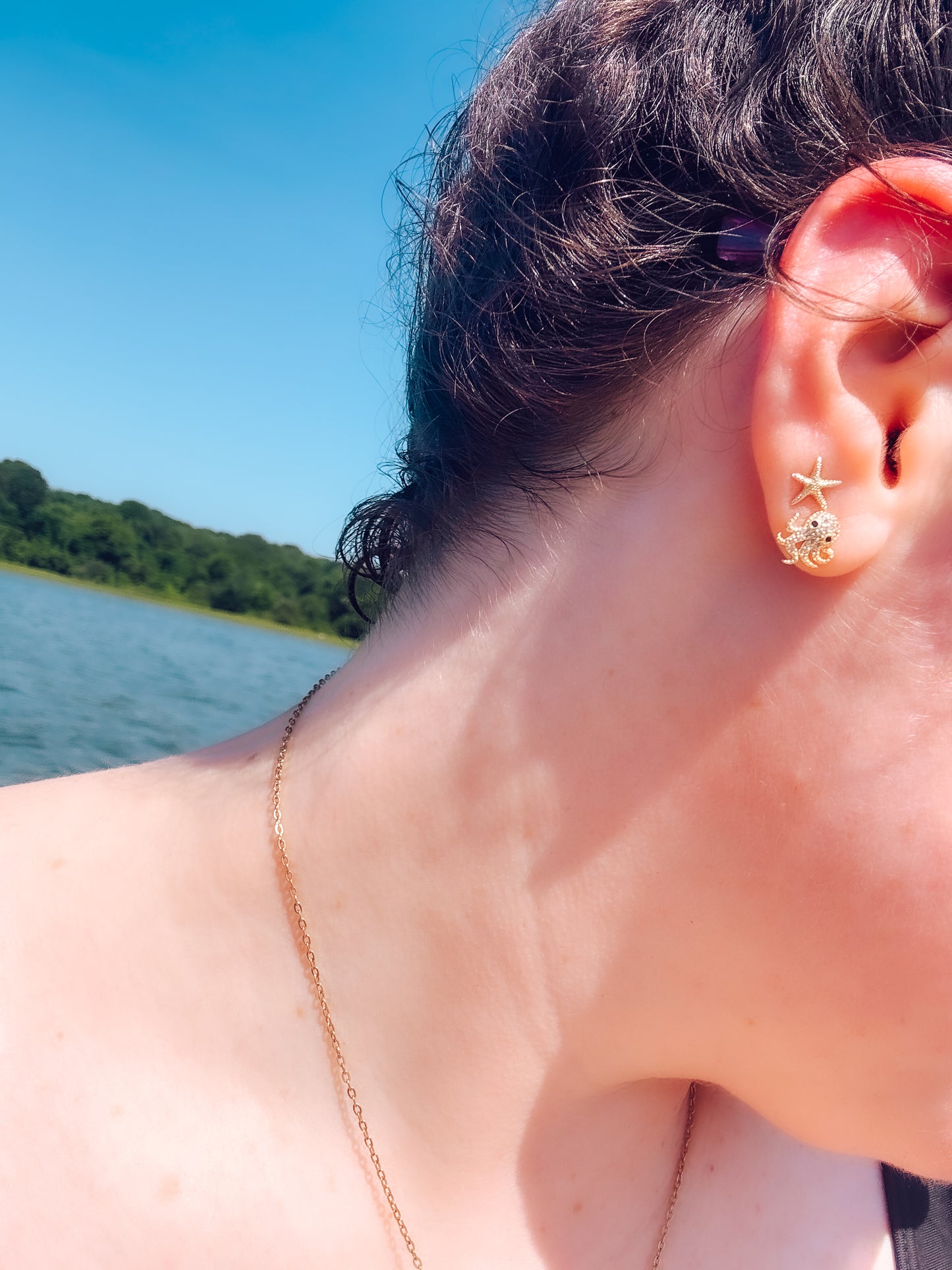 A close-up lifestyle shot of a person's ear wearing a gold starfish stud earring, with a delicate gold chain necklace and a lake background under a blue sky.