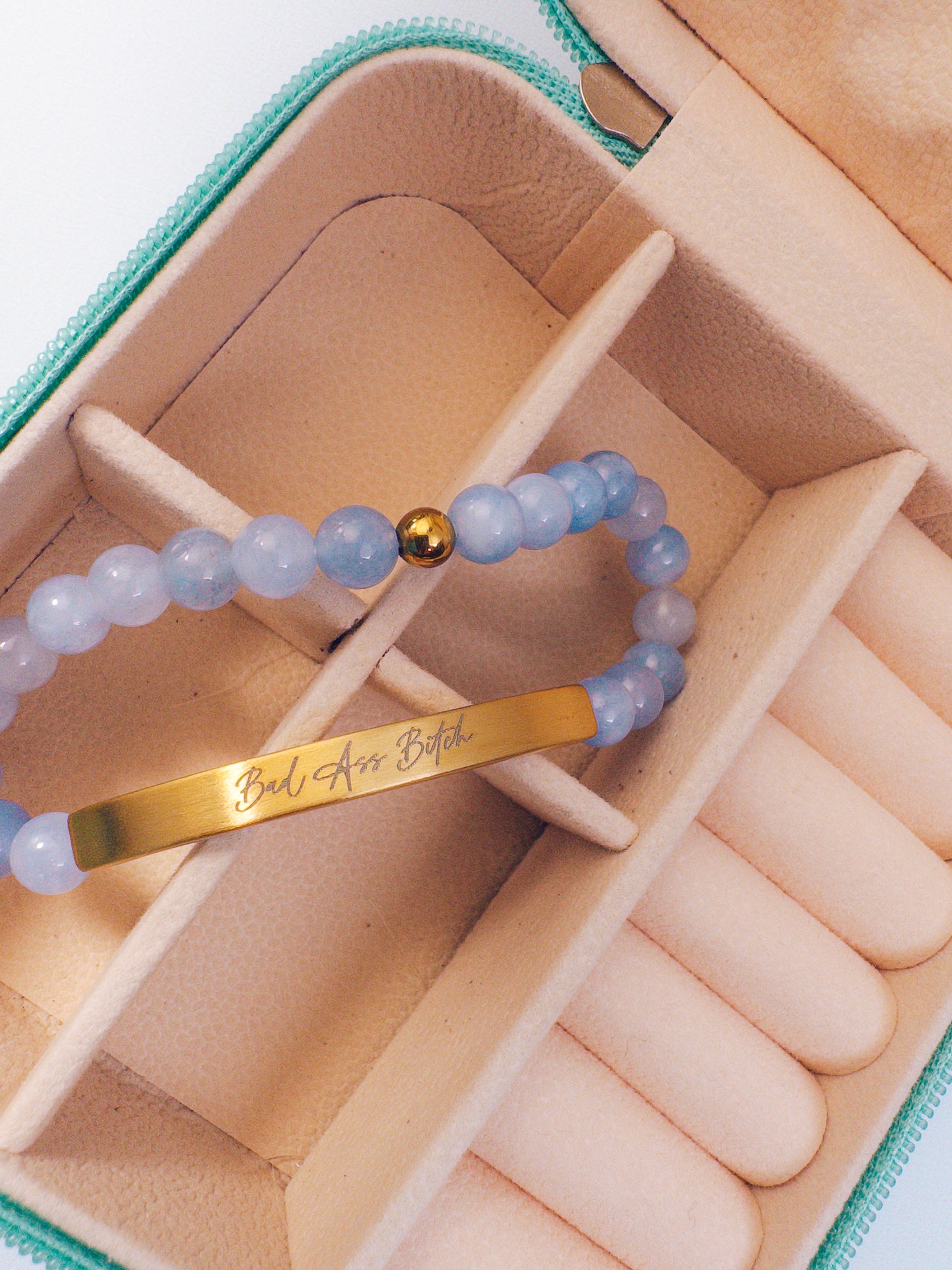 A close-up of a beaded bracelet with light blue translucent beads, a small gold ball accent, and a gold bar engraved with the phrase 'Bad Ass Bitch', displayed inside a beige jewelry organizer box.