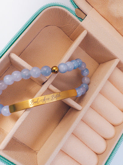 A close-up of a beaded bracelet with light blue translucent beads, a small gold ball accent, and a gold bar engraved with the phrase 'Bad Ass Bitch', displayed inside a beige jewelry organizer box.