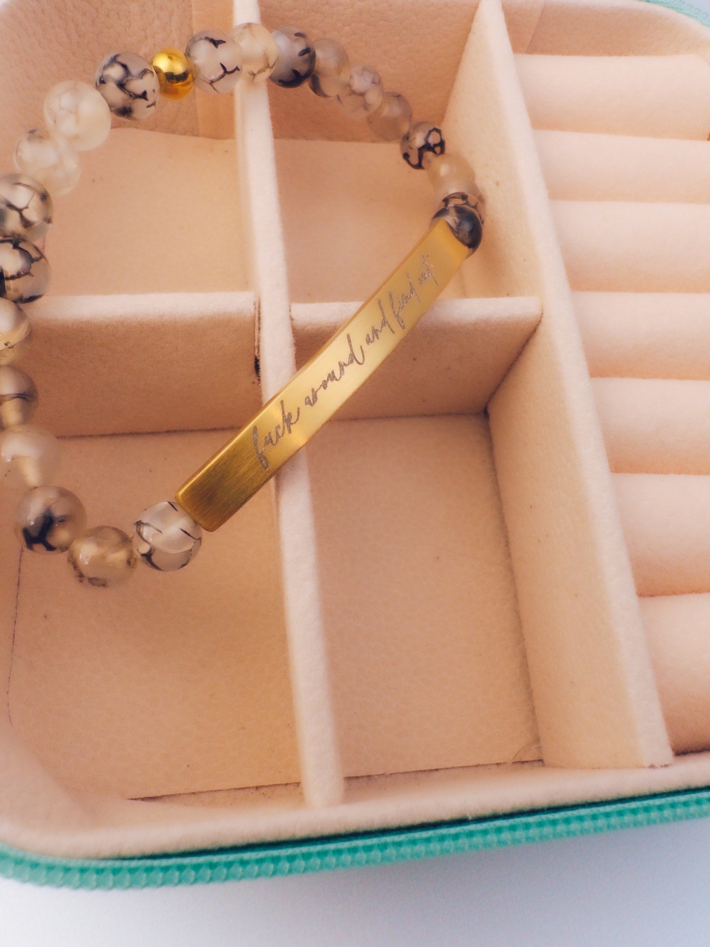 A beaded bracelet with a gold bar engraved with 'fuck around and find out' resting inside a beige jewelry organizer box.