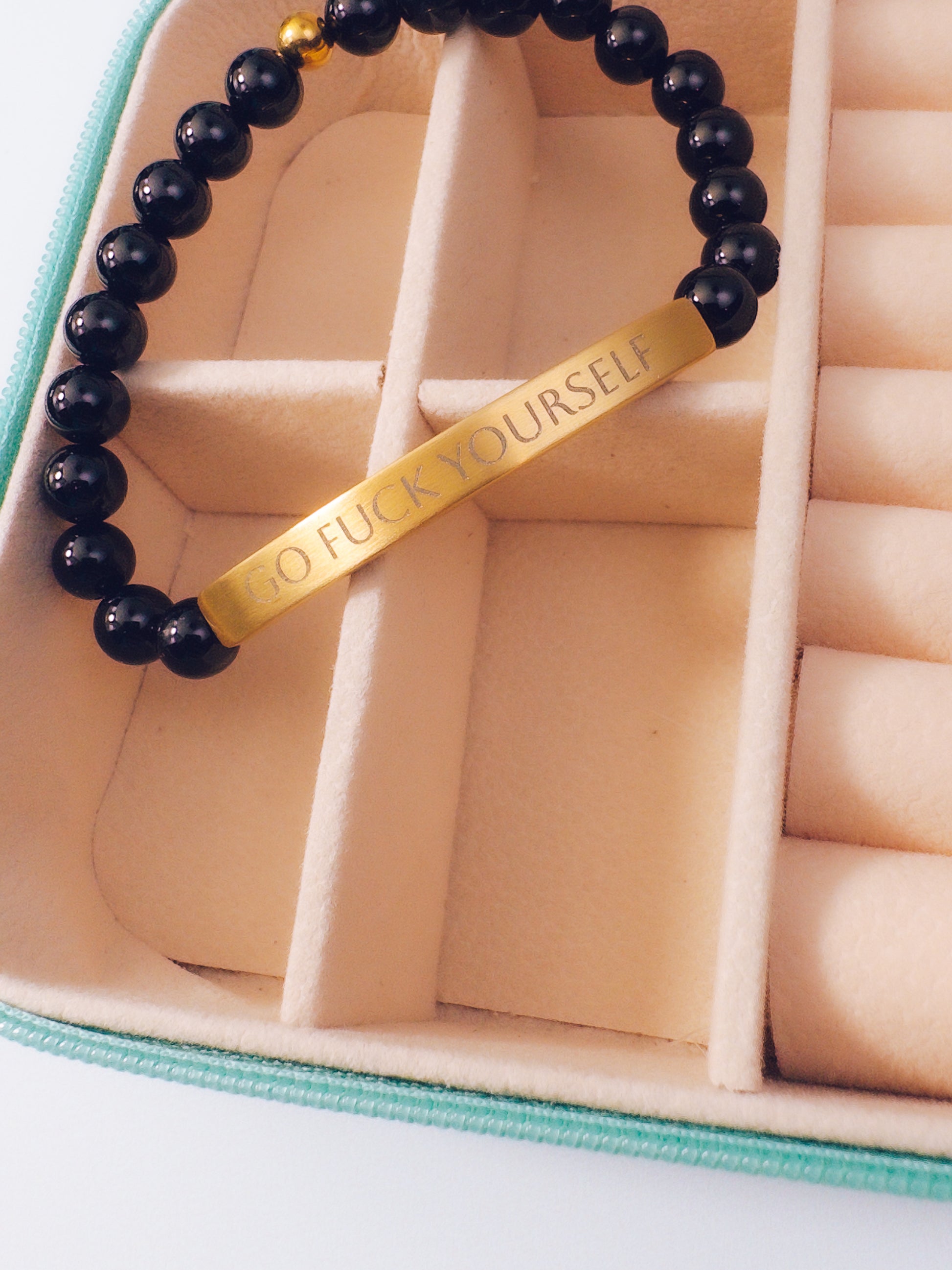 A beaded bracelet with glossy black round beads and a gold bar engraved with the phrase 'GO FUCK YOURSELF', displayed inside a beige compartmentalized jewelry box.