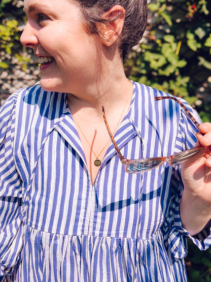 Woman wearing a blue and white striped dress holding sunglasses, with greenery in the background