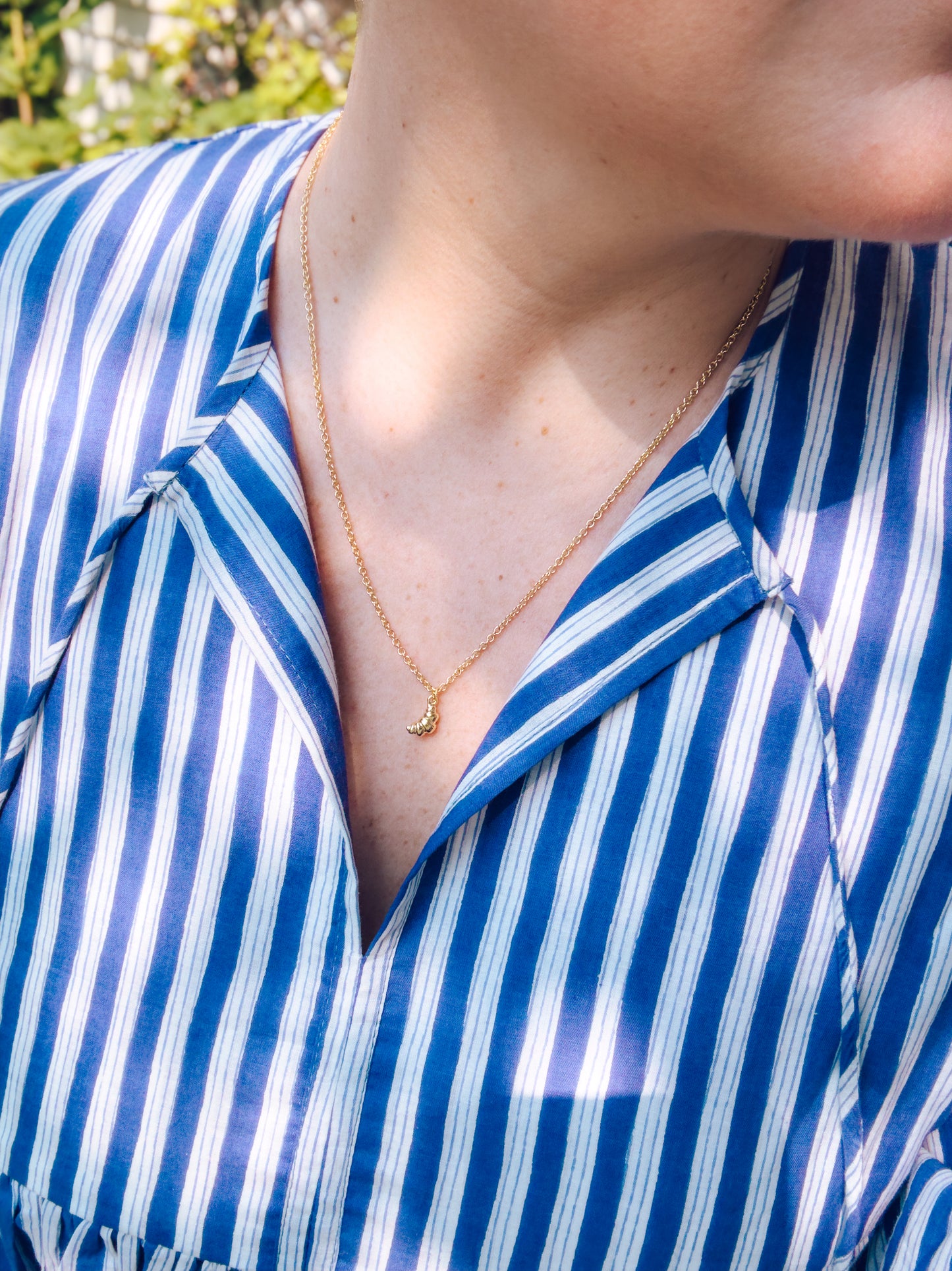 Person wearing a gold necklace with a small pendant against a blue and white striped shirt.
