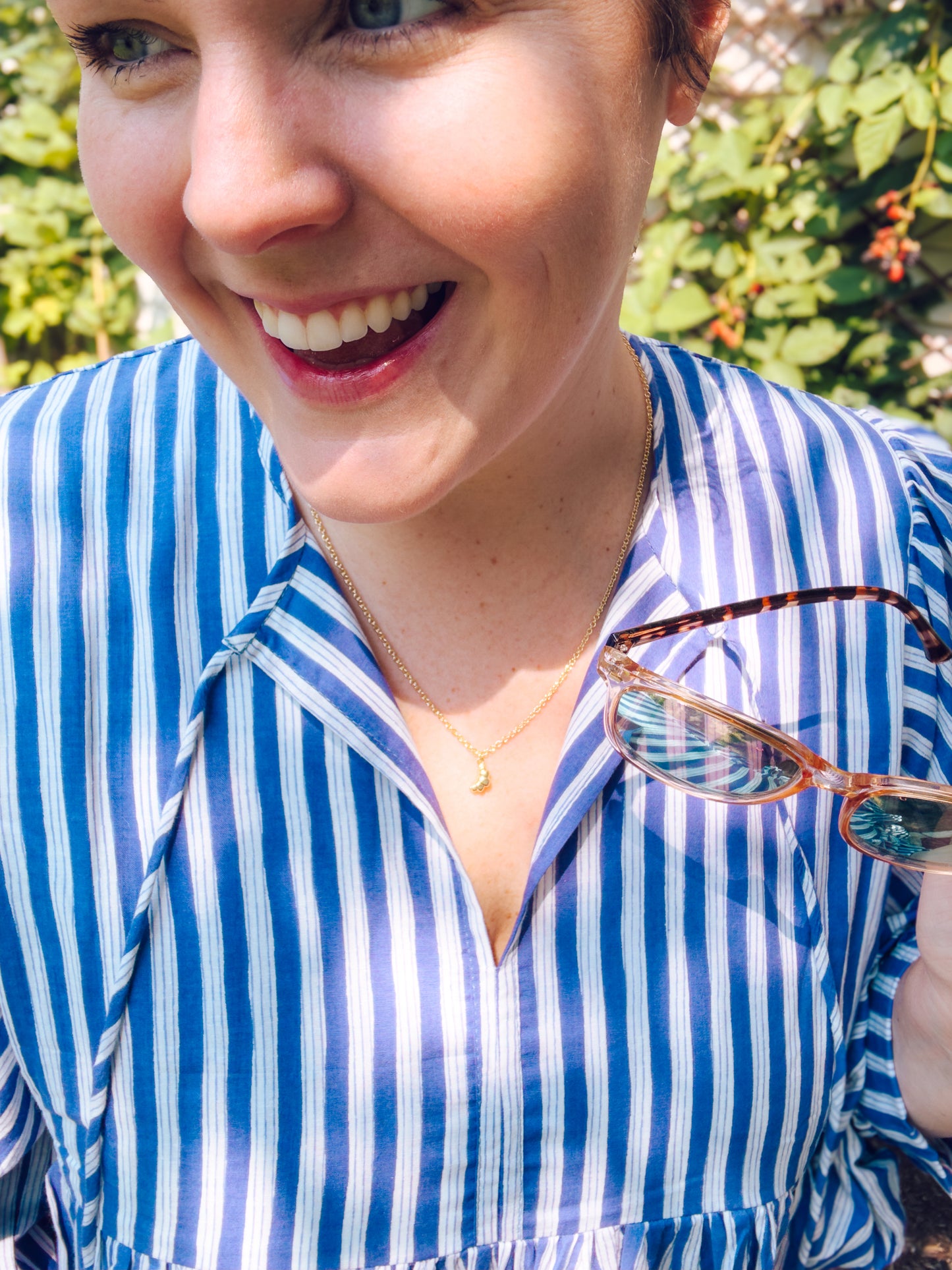 Person wearing a blue and white striped shirt holding sunglasses, with greenery in the background