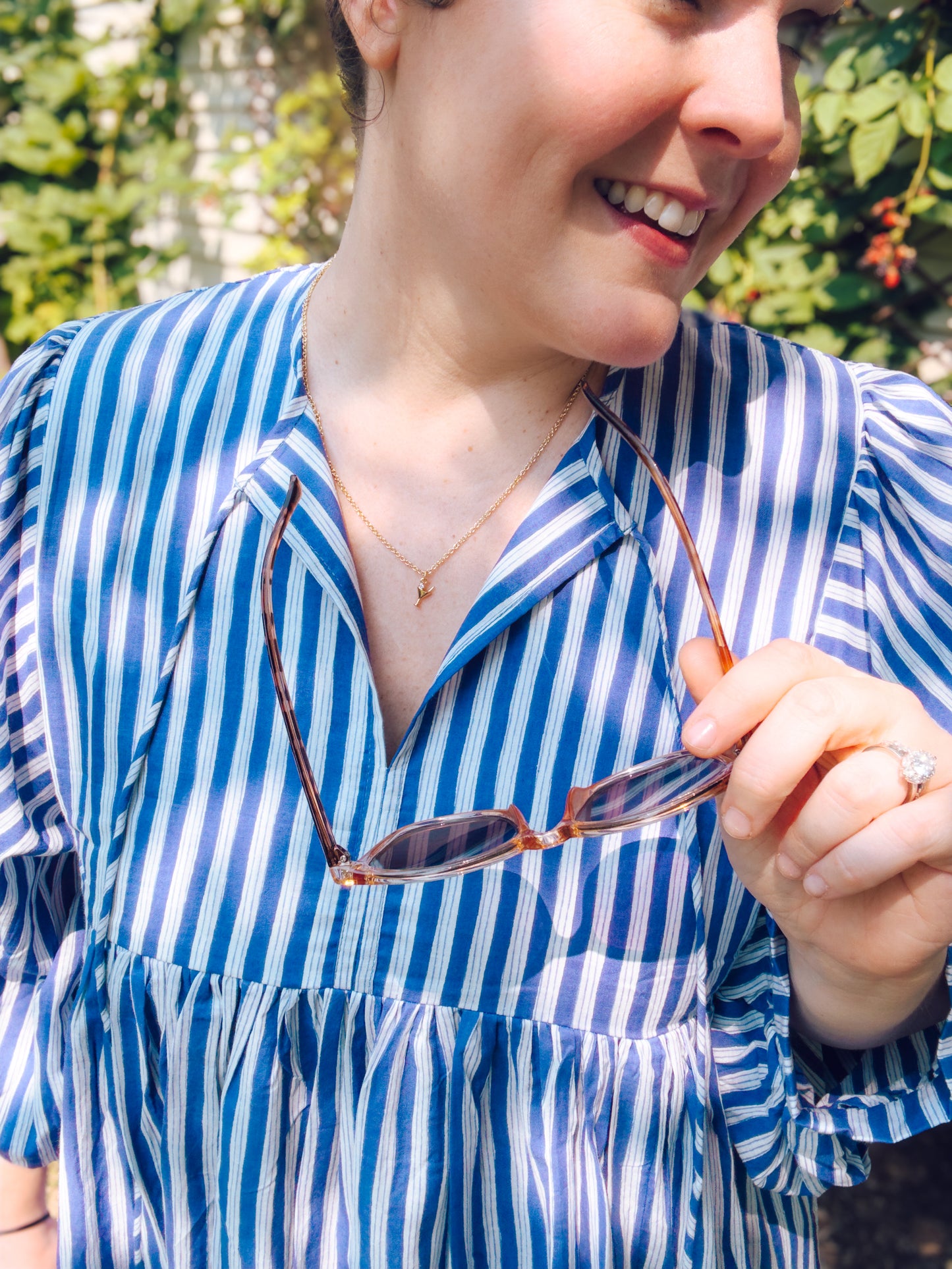 Person wearing a blue and white striped shirt holding sunglasses, with a blurred natural background