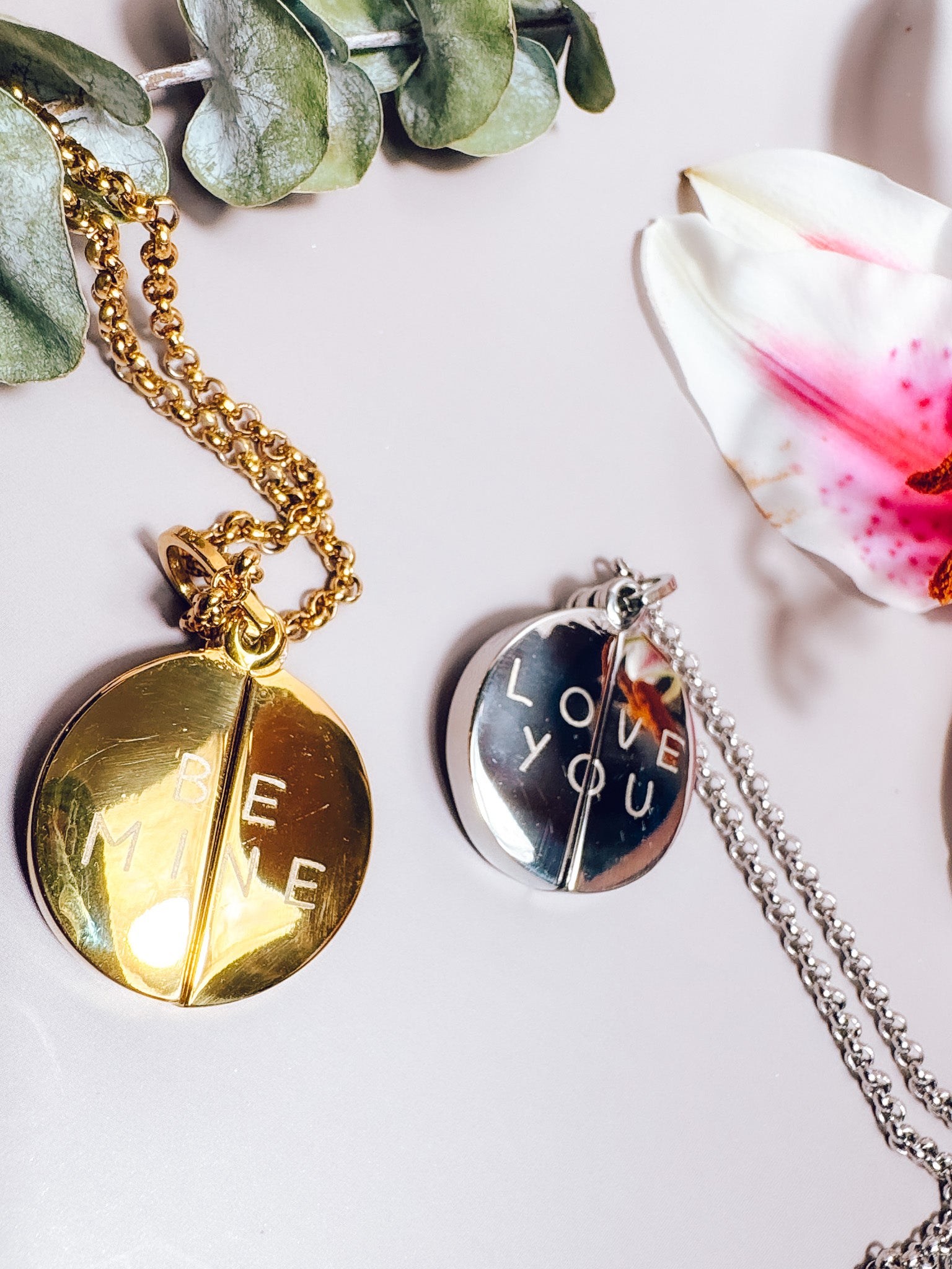 Two necklaces with round pendants on a light background with greenery and a flower.