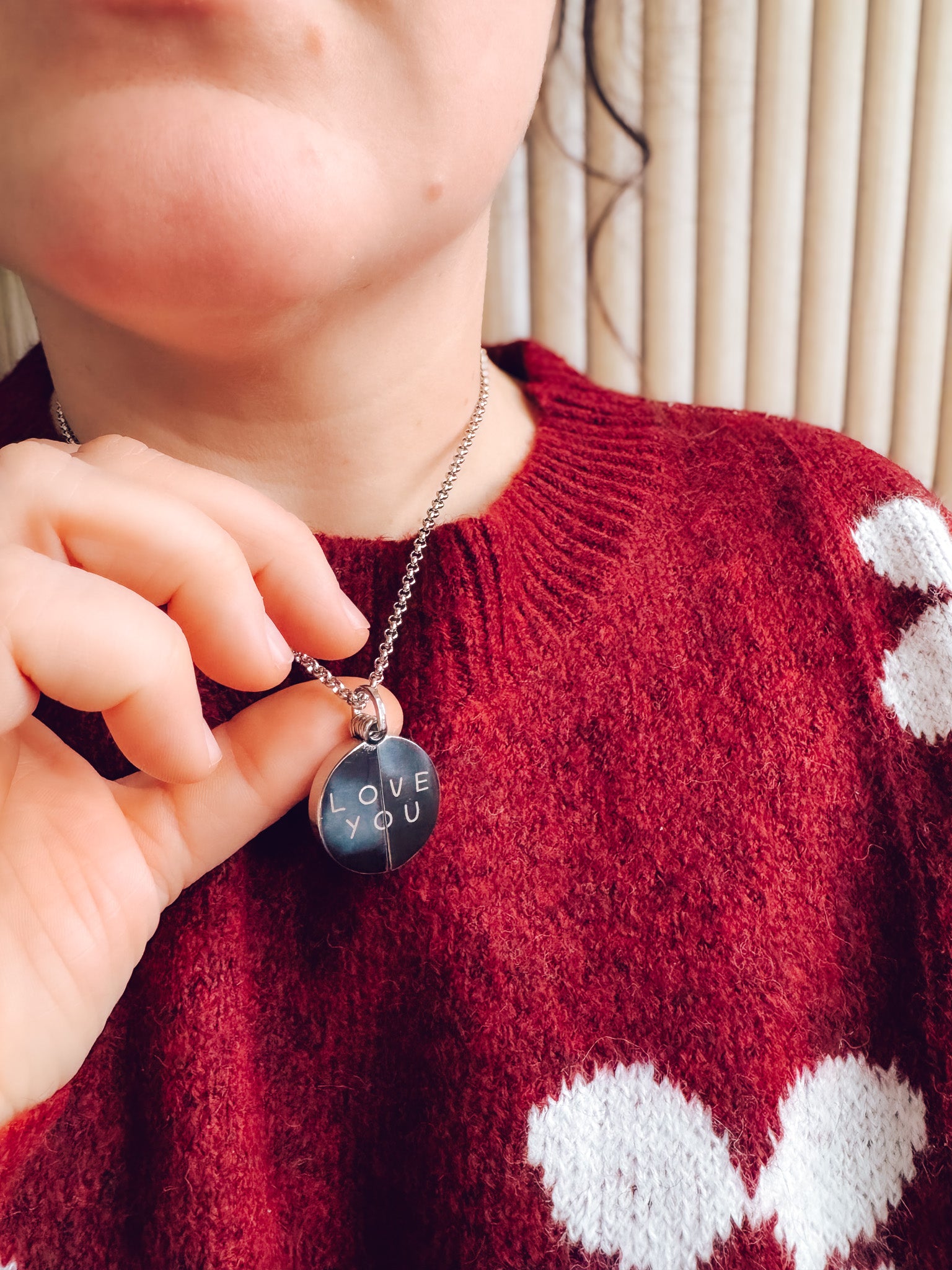 Person wearing a red sweater with white heart patterns, holding a necklace with a 'LOVE YOU' pendant.